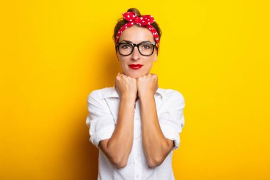 Genç bir kadın ellerini çenesinde tutuyor, gözlük takıyor ve sarı bir arka planda saç bandı takıyor. Pankart.