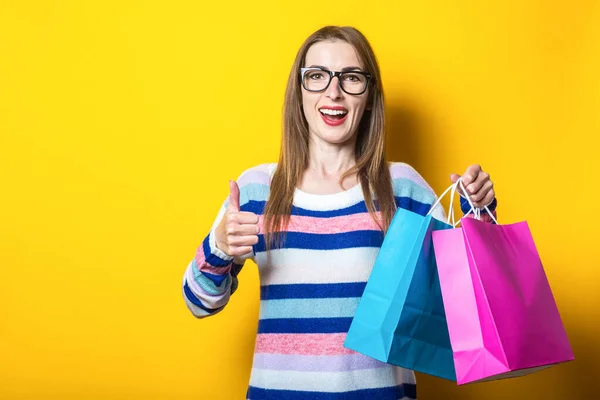 Young woman smiles and makes a gesture all is well, holds and shopping bags with purchases on a yellow background. Banner.