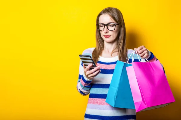 Young woman looks into the phone and holds shopping bags with purchases on a yellow background. Banner.