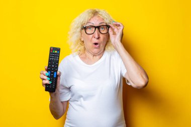 Surprised blonde old woman with glasses holding remote control on yellow background clipart