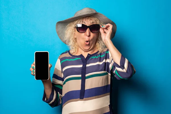 Surprised old woman in glasses and hat holding phone on blue background