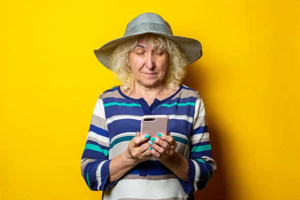 Old woman in hat looking at phone on yellow background