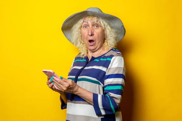 Surprised old woman in hat holding phone on yellow background