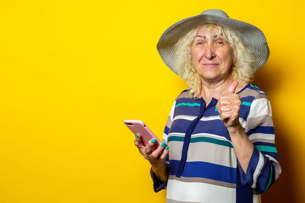 Surprised shocked old woman in hat holding phone shows gesture class on yellow background