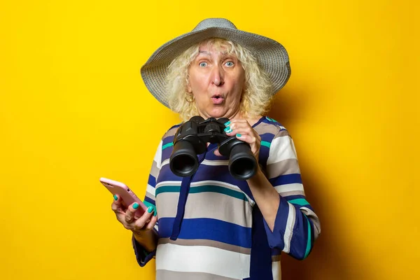 Surprised shocked old woman in hat holding phone and binoculars on yellow background
