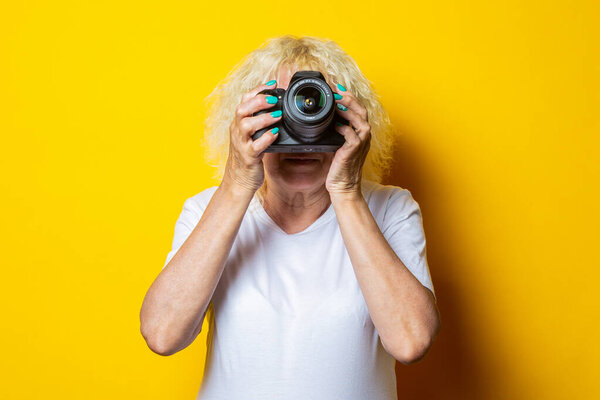 Smiling blonde old woman in white t-shirt holding a camera and taking pictures on yellow background