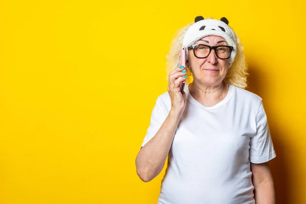 Smiling blond old woman in sleep mask holding phone on yellow background