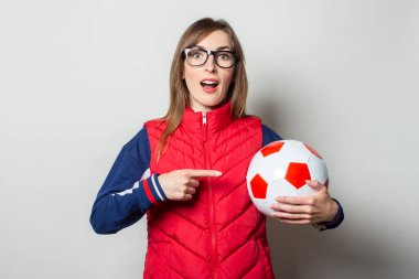 Kırmızı yelekli genç bir kadın elinde bir futbol topu tutuyor ve arka planda parmağıyla topu işaret ediyor..