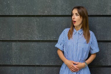 Elbiseli, şaşırmış yüzlü genç bir kadın taş bir duvarın arkasına bakıyor..