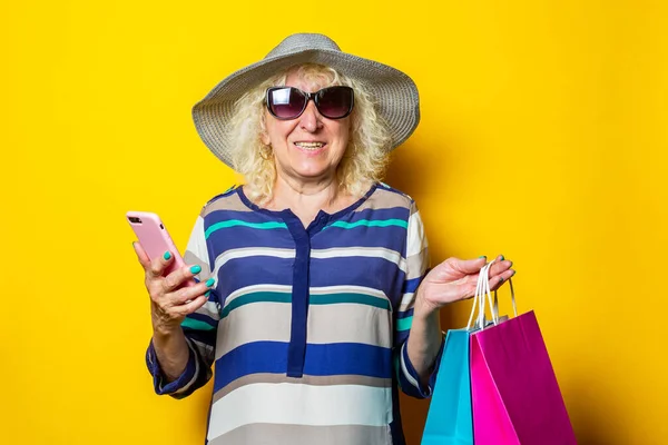Old woman in hat and glasses holds shopping bags with purchases and phone on yellow background.
