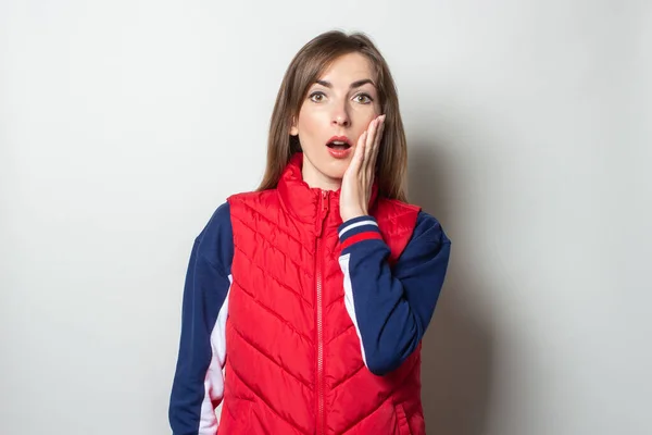 Young woman with a surprised face in a red vest on a light background.
