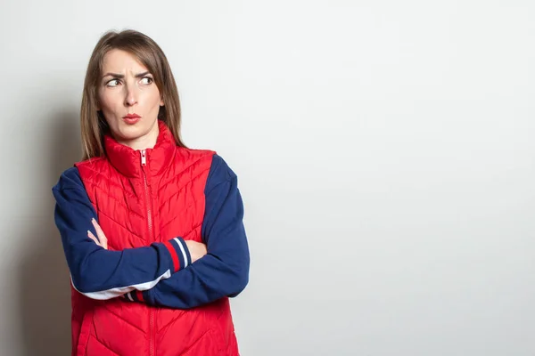 Young woman with a serious face looks to the side in a red vest on a light background.