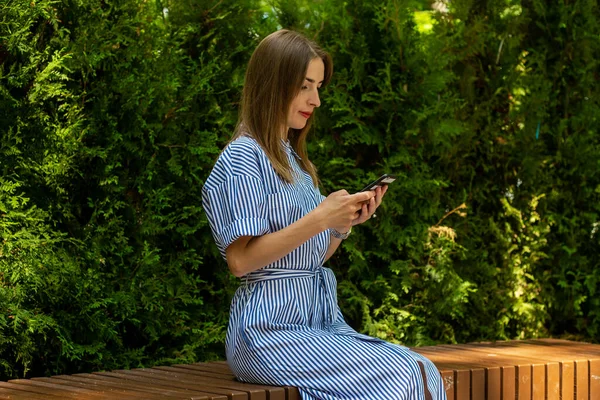 드레스를 우정적 여자가 벤치에 공원에서 자신의 핸드폰을 있습니다 — 스톡 사진