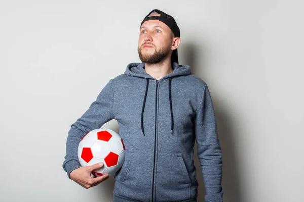 Young man in a cap and hoodie holds a soccer ball and looks to the side on a light background.