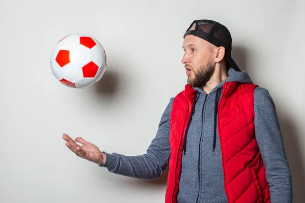 Young man in a cap, hoodie and vest throws up a soccer ball on a light background.