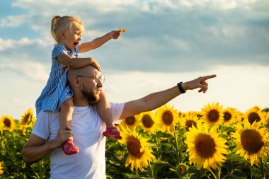 Gözlüklü genç bir adam bir çocuğu omuzlarında tutuyor ve bir ayçiçeği tarlasında bir şeye dokunuyor..