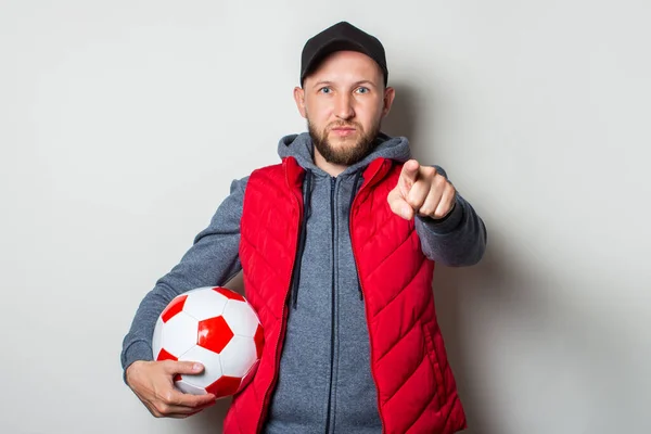 Young man in a cap, hoodie and vest holds a soccer ball and points his finger at the viewer on a light background. You are next gesture.