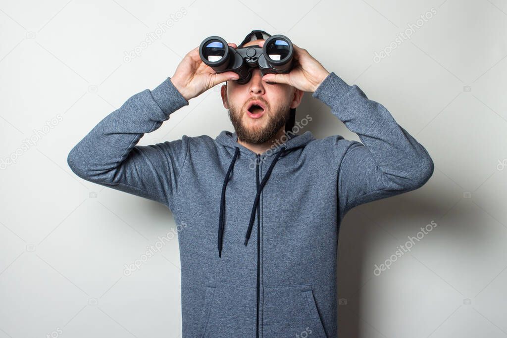 Young man with a surprised face in a hoodie and a cap looks through binoculars on a light background.