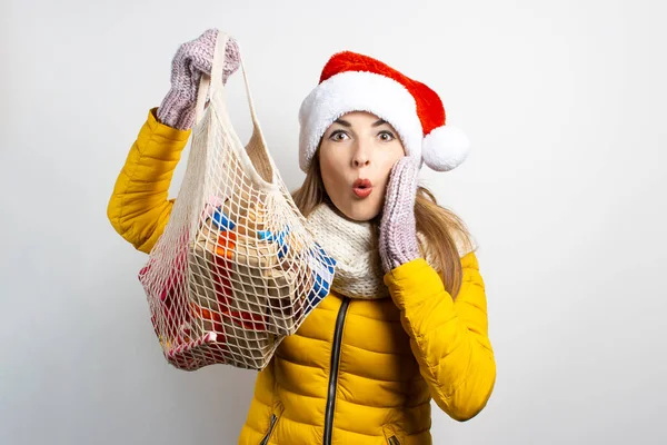 Young woman with a surprised face in Santa Claus hat is holding a shopping bag with gifts on a light background. Concept of the winter holidays, Christmas, gift shopping. Mood, emotions, surprise.