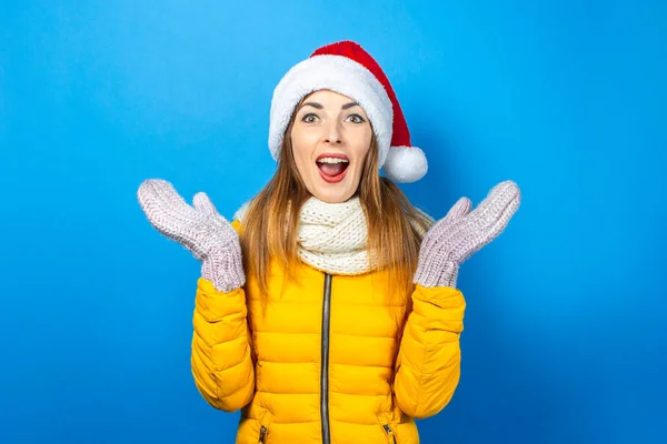 Young woman with a surprised face in a yellow jacket and a hat of Santa Claus on a blue background. Concept of the winter holidays, Christmas, New Year, surprise, shock. Banner.