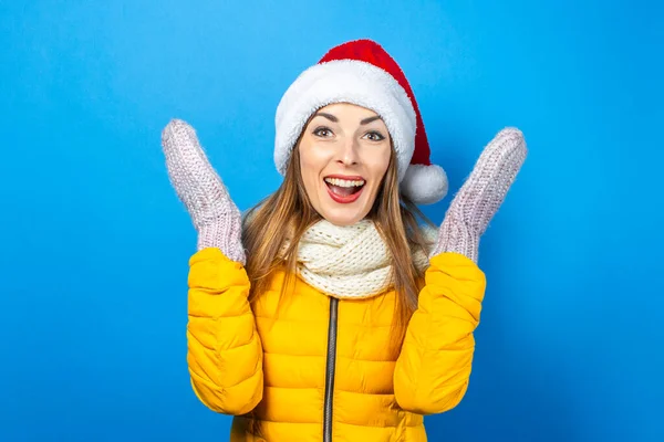 Young woman with a surprised face in a yellow jacket and a hat of Santa Claus on a blue background. Concept of the winter holidays, Christmas, New Year, surprise, shock. Banner.