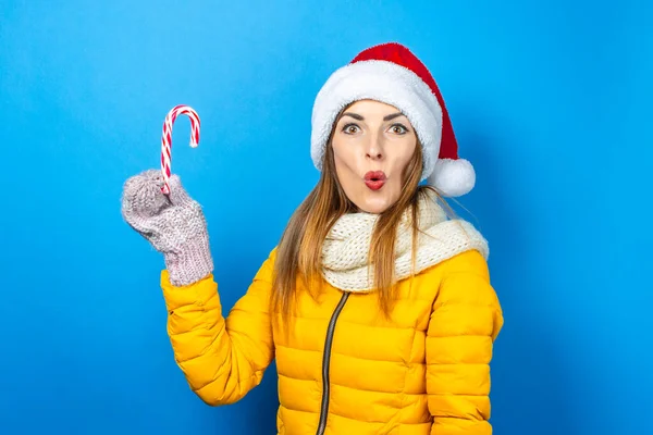 Young woman with a surprised face in a yellow jacket and hat of Santa Claus holds a candy cane on a blue background. Concept of the winter holidays, Christmas, New Year, surprise, shock. Banner.