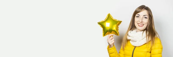 Woman with smile in a yellow jacket and a scarf holds a star air balloon on a white background. Concept of a holiday, celebration, party, new year. Banner.