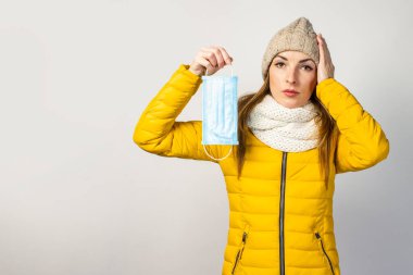Sarı ceketli ve şapkalı genç bir kız, tıbbi bir maske tutuyor, hasta ve elini hafif bir arka planda kafasına dayıyor. Soğuk algınlığı, virüs, bulaşıcı hastalıklar. 