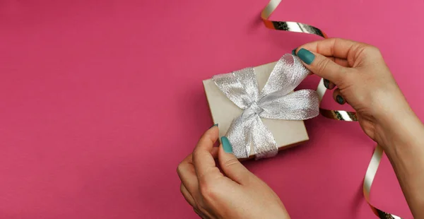 Gift box with a silver bow and a girl holds in her hands and unties a bow on a pink background for the feast day of St. Valentine