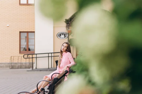 Egy fiatal, gyönyörű lány rózsaszín ruhában sétálgat a városban. Fénykép közelről - és Általános terv. Lány ül a padon. — Stock Fotó