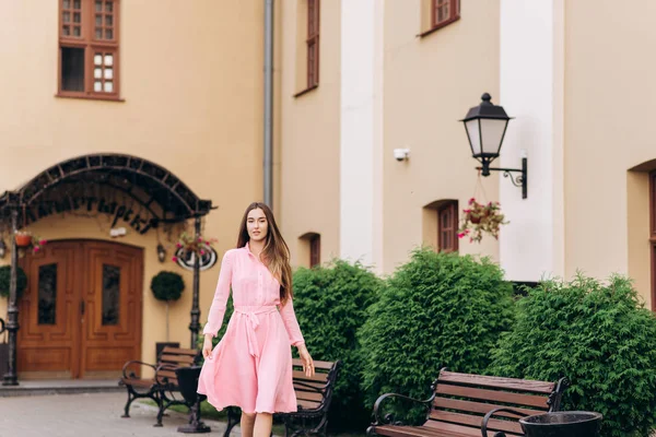 Egy fiatal, gyönyörű lány rózsaszín ruhában sétálgat a városban. Fénykép közelről - és Általános terv. Lány ül a padon. — Stock Fotó