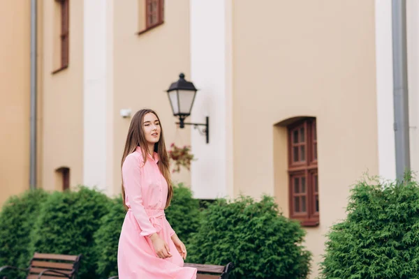 Uma jovem, linda menina em um vestido rosa caminha pela cidade. Foto de perto - para cima e plano geral. Menina sentada em um banco . — Fotografia de Stock