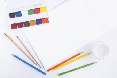 Düz parlak çok renkli suluboya boyalar, boyama için fırçalar, yıkama fırçaları için bir konteyner, renkli ahşap kalemler ve metin veya boş sayfaları ile açık bir albüm ile güzel kompozisyon yatıyordu