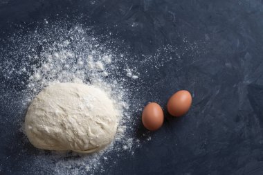Metin ve diğer tasarım öğeleri için boş bir yer ile dokulu koyu mavi rustik arka plan üzerinde buğday unu tozu ile ham pişirme hamuru ve iki kahverengi yumurta bir top düz yatıyordu