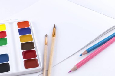 Parlak suluboya boyaların yan görünümü, çizim için iki fırça, iki renkli kalem ve beyaz bir masada boş sayfalarla çizmek için açık bir albüm