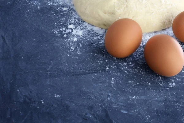 Çiğ ekmek hamuru bir top, metin ve diğer tasarım öğeleri için boş alanlar ile mavi dokulu beton arka plan üzerinde resmin köşesinde beyaz buğday unu ve üç kahverengi yumurta serpilir — Stok fotoğraf