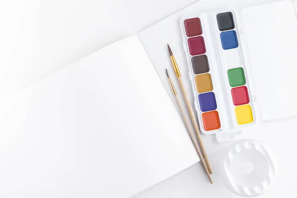 Flat lay design composition of bright watercolor paints, two brushes, a container for washing brushes and an open album for drawing with blank pages for text or a picture on a white background