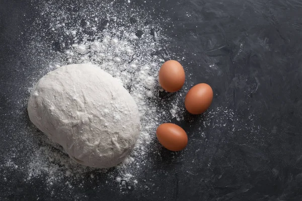 Pişirme, pişirme, ekmek, pasta ve kek için malzemelerin bir grup üzerinde üst görünümü. Üç kahverengi tavuk yumurtası, çiğ hamur pişirme bir top ve siyah dokulu arka plan üzerine beyaz un serpilir — Stok fotoğraf