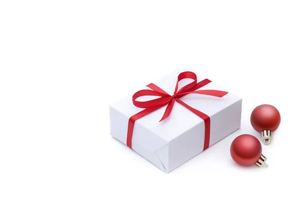 Boční pohled na bílou krabičku dárků svázaný červenou satínovou jasnou stuhou s lukem a dvěma červenými sváteční vánoční míčky s prázdným místem pro text izolovaný na bílém pozadí — Stock fotografie