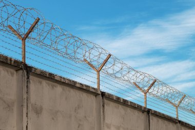 Açık mavi gökyüzünün karşısındaki gri beton bir çitin üzerindeki çelik süslemeli dikenli tel. Metin için boş bir yeri olan bulutlar, iki zıt şeyin sembolü..