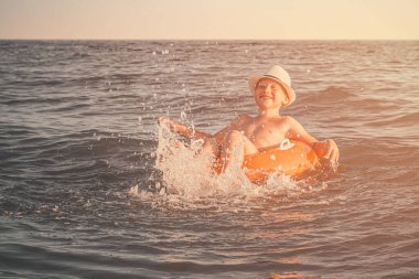 Turuncu yüzme çemberinde beyaz şapkalı neşeli beyaz bir çocuk bacaklarını denize atıp gülümsüyor. Bir çocuk gün batımında otelde yüzer ve eğlenir. Tatiller, seyahat, turizm konsepti.