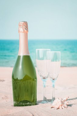 Mavi gökyüzü ve okyanusun arka planına karşı beyaz kumda iki bardak ve bir şişe şampanya.