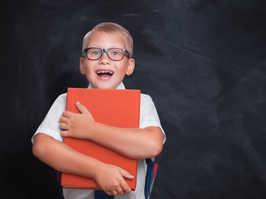 Beyaz tişörtlü, gözlüklü, beyaz tenli küçük beyaz bir çocuk kırmızı bir kitaba sarılıyor..