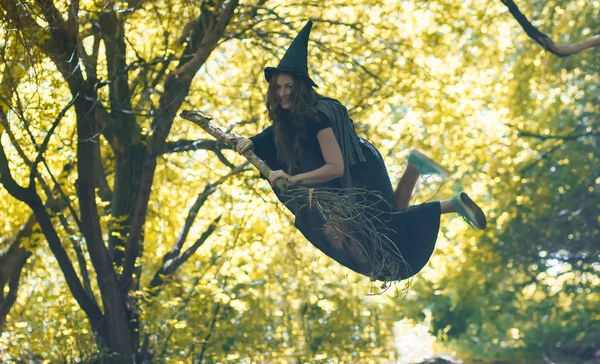女巫骑在扫帚上飞行 在秋天的森林里 一个戴着女巫帽的漂亮姑娘 — 图库照片