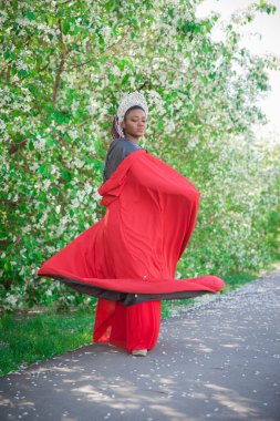 Kraliçe tacı ve kraliyet pelerini. Çiçek açan bir bahçenin arka planında güzel bir kız. Kırmızılı Afrikalı Amerikalı