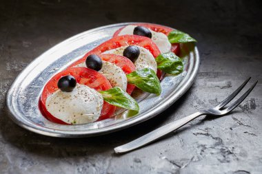 Caprese salatası. Domates ve mozzarella peyniri dilimleri, fesleğen ve zeytin bir plaka üzerinde var. Koyu arka plan.