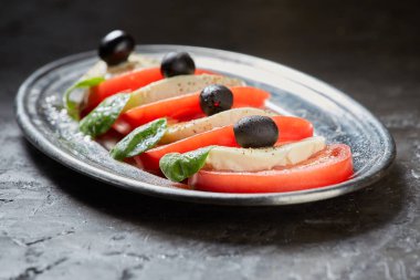 Caprese salatası. Domates ve mozzarella peyniri dilimleri, fesleğen ve zeytin bir plaka üzerinde var. Koyu arka plan.