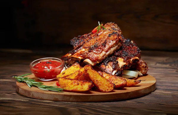 Fried ribs with rosemary, potatoes rustic, onion, sauce on wooden round Board. Dark background. Place for text, copyspace