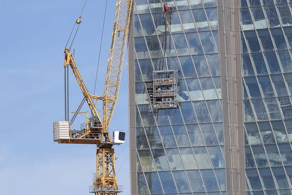 Tower crane on the background of the glass building. The installation of glass at height. The background of blue sky