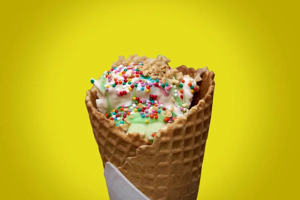 white ice cream in a waffle cone with colored sprinkles and pieces of nuts yellow background